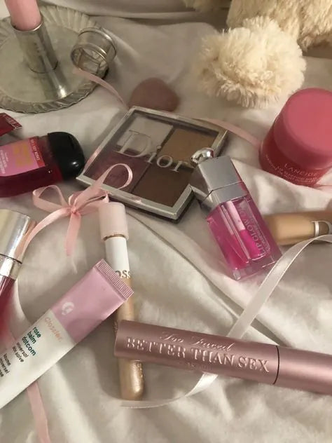 Luxury makeup set including pink lipstick, eyeshadow palette, lip gloss, lip liner, and mascara on a white bedspread.